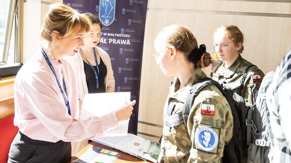 Zdjęcie przedstawia kobietę z folderem, przed którą stoi uczennica w mundurze